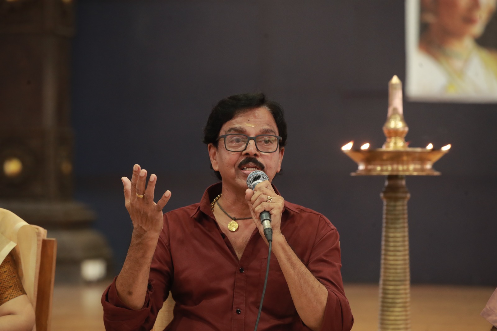 Guru V. S. Janardhanan speaking into a microphone beside a lit brass nilavilakku lamp